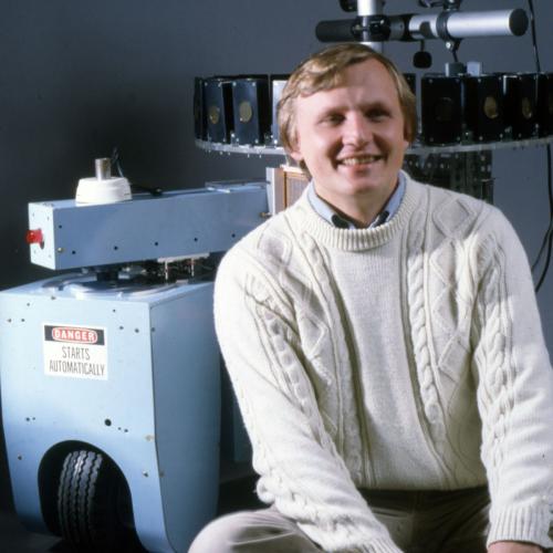 An image of a man in a white sweater sitting in front of a blue robot, which has a sonar ring attached.