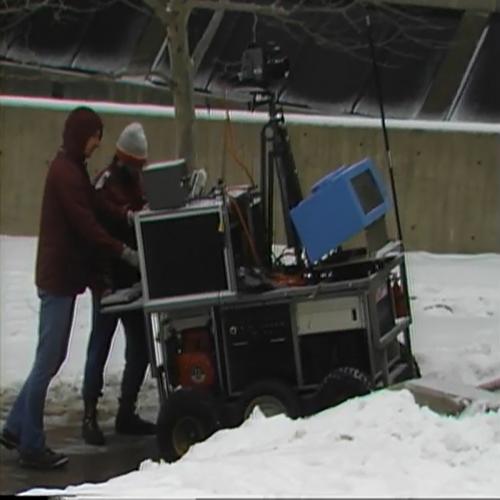 An image of two men walking behind a six-wheeled robot moving on a sidewalk during winter. A camera is mounted on a tripod on top of the robot and a blue box-like sensor sits on the front