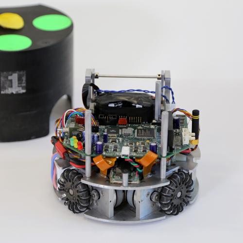 An image of a small, wheeled, circular robot with its inner circuitry displayed. A black plastic covering for the robot is in the background.